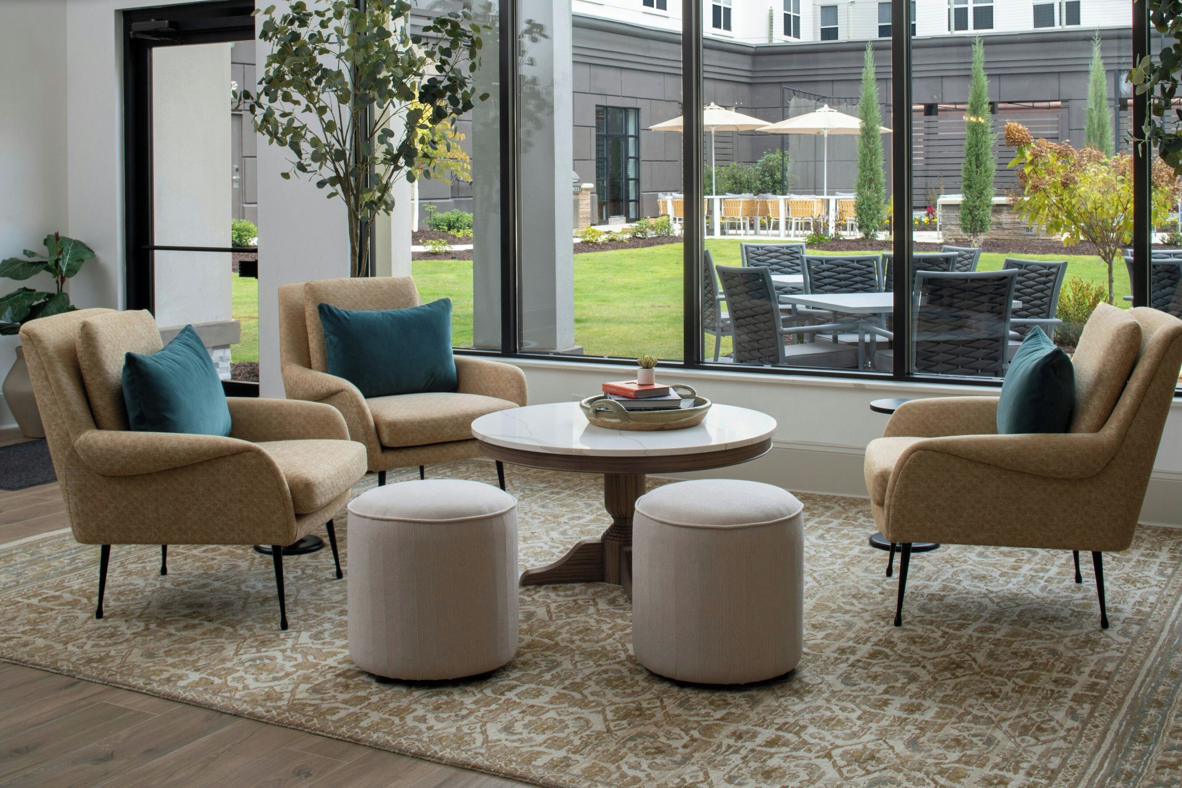 A modern living room with beige armchairs, a round coffee table, and ottomans. Large windows offer a view of the outdoor patio area with outdoor furniture and a garden.