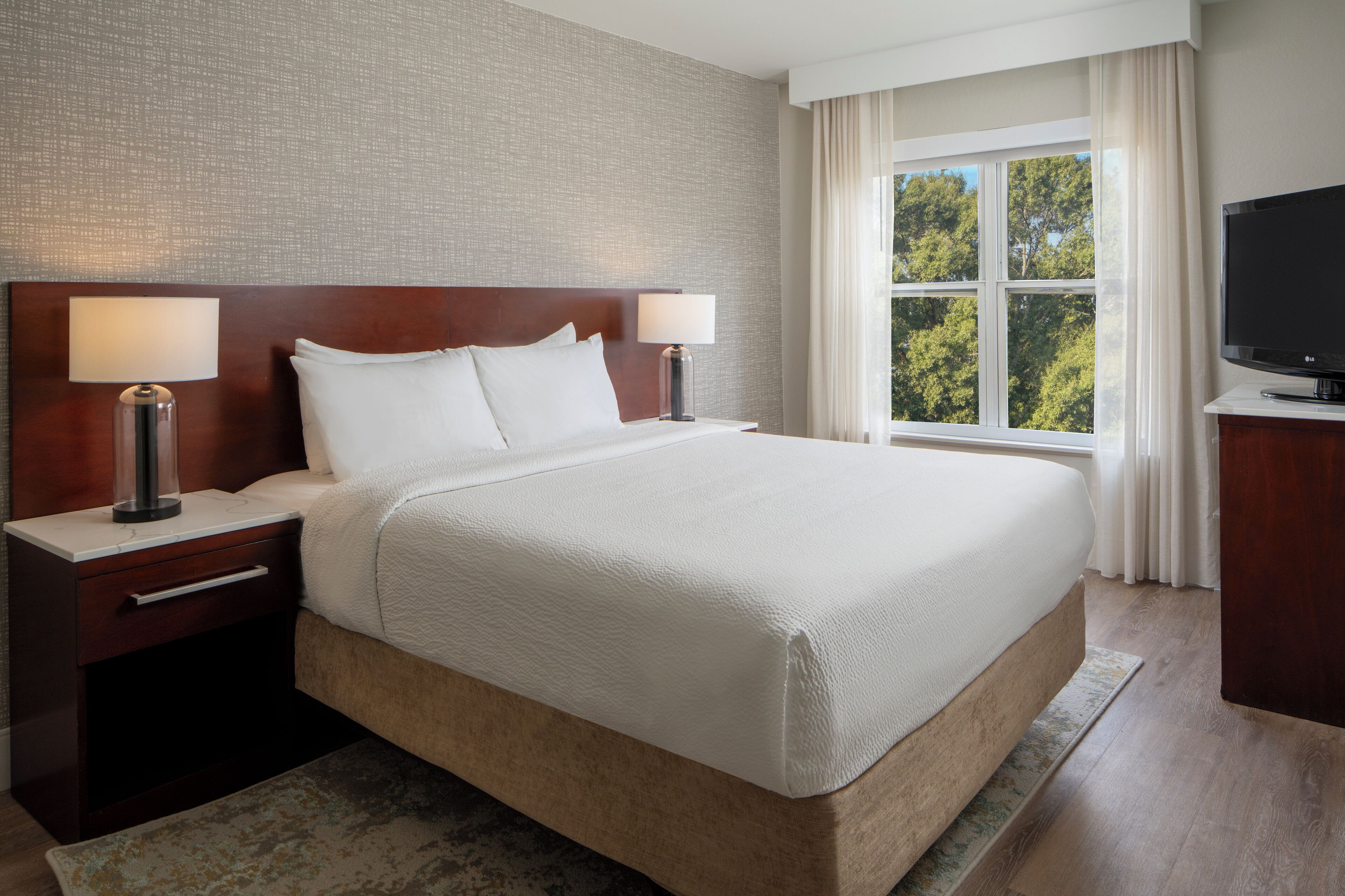 A hotel room with a large bed, two bedside tables with lamps, a flat-screen TV, and a window with curtains.