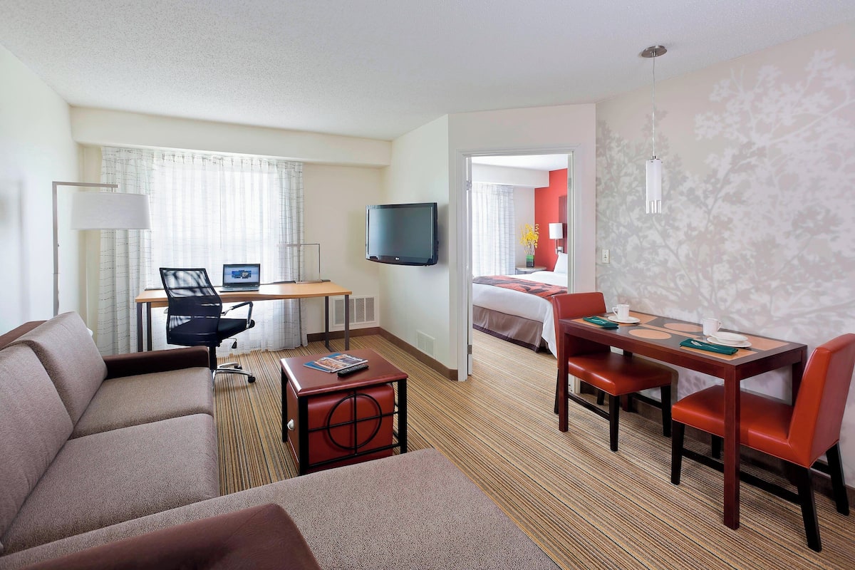 A hotel room with a sofa, a dining table with chairs, a TV mounted on the wall, and a separate bedroom area.