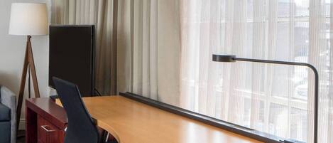 A hotel room with a large desk, a chair, a TV, a lamp, and a view of the city through sheer curtains.