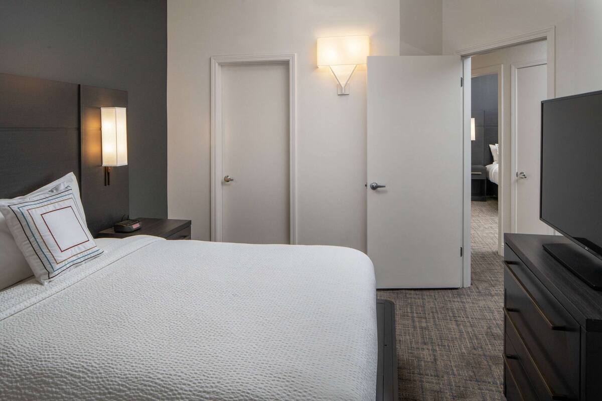 Una habitación de hotel con una cama grande, televisor, mesita de noche y lámpara de pared.