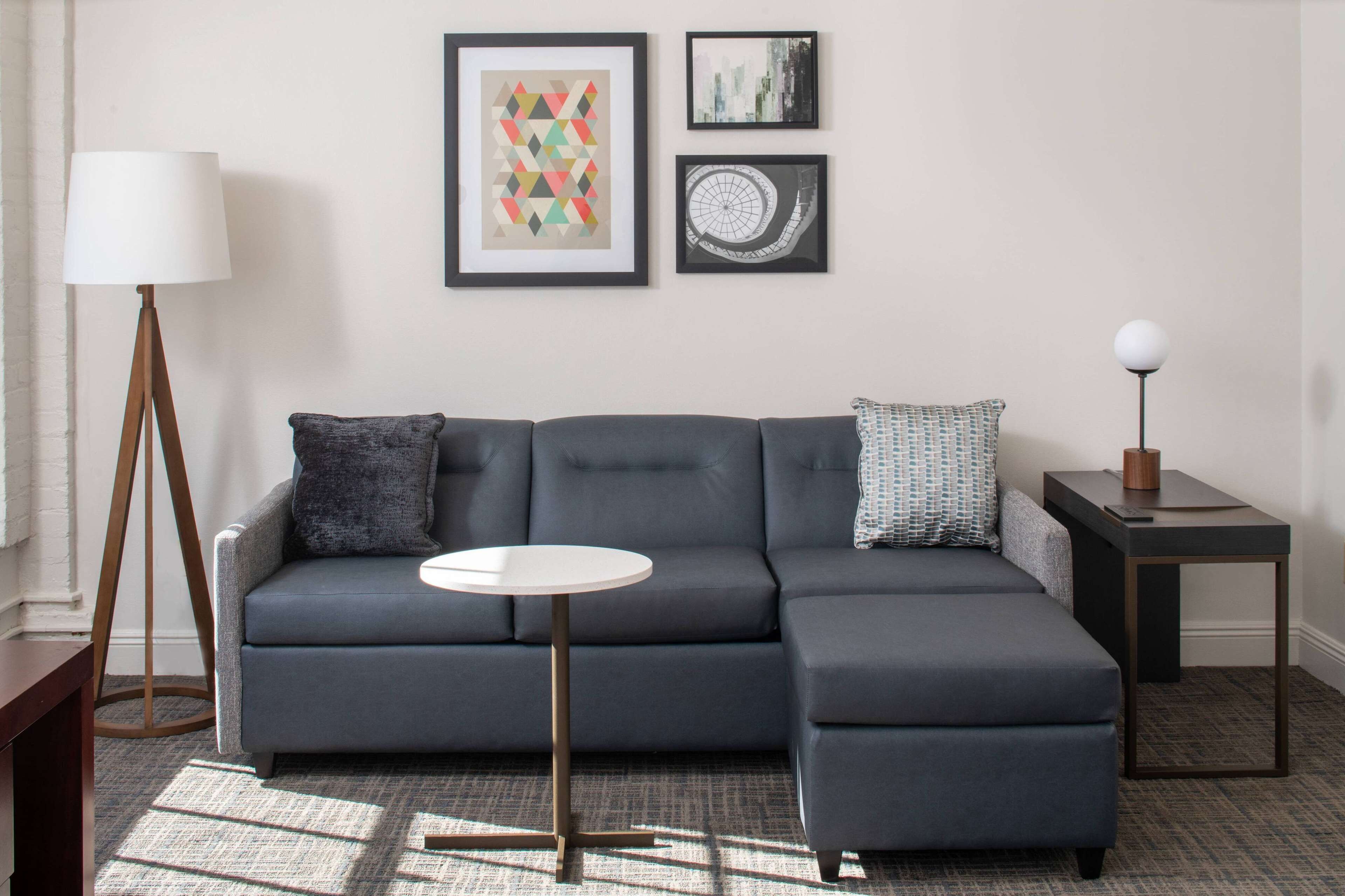 A modern living room with a grey sectional sofa, a round coffee table, and a small side table with a lamp. There are three framed artworks on the wall.