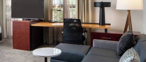Quarto de hotel com sofá, mesa pequena, televisão, escrivaninha e luminária.