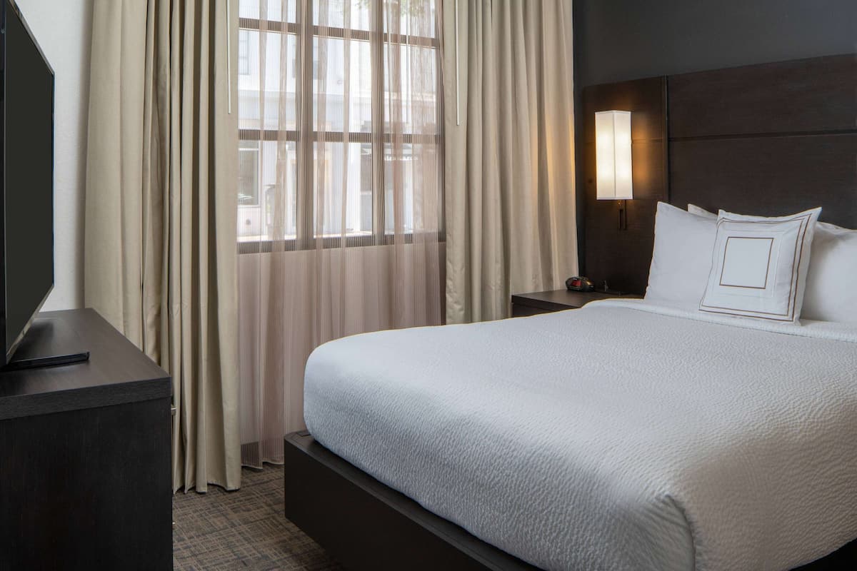 Una habitación de hotel con una cama grande, un televisor sobre un mueble, una mesita de noche, una lámpara y cortinas traslúcidas.