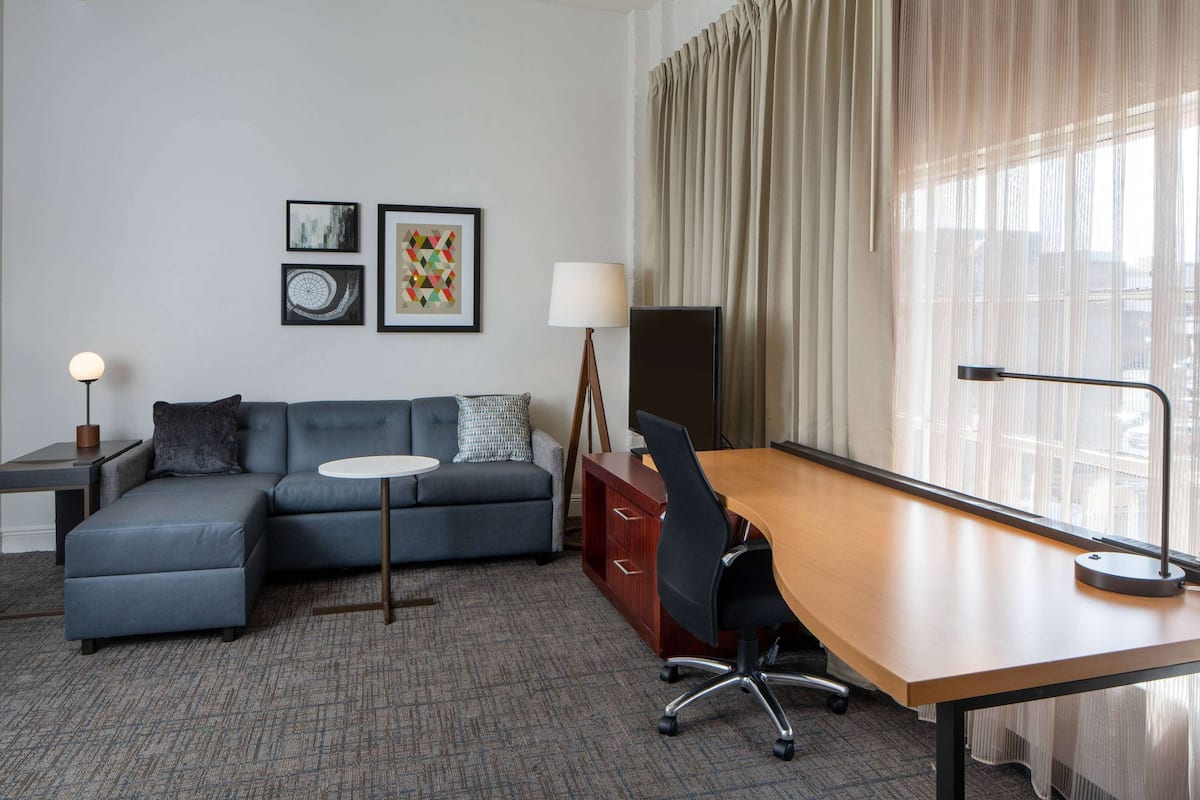 Una habitación de hotel moderna con un sofá seccional gris, un escritorio de madera, una silla de oficina negra, una mesita redonda pequeña, una lámpara, un televisor y obras de arte en la pared.