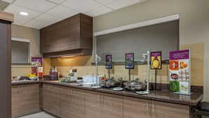 A hotel kitchen with wooden cabinets, a granite countertop, and various breakfast items displayed on the wall.