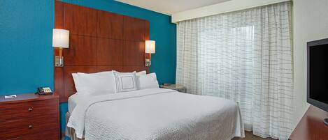 Una habitación de hotel con una cama grande, cabecera de madera, dos lámparas de noche, mesitas de noche, un televisor y una ventana con cortinas.