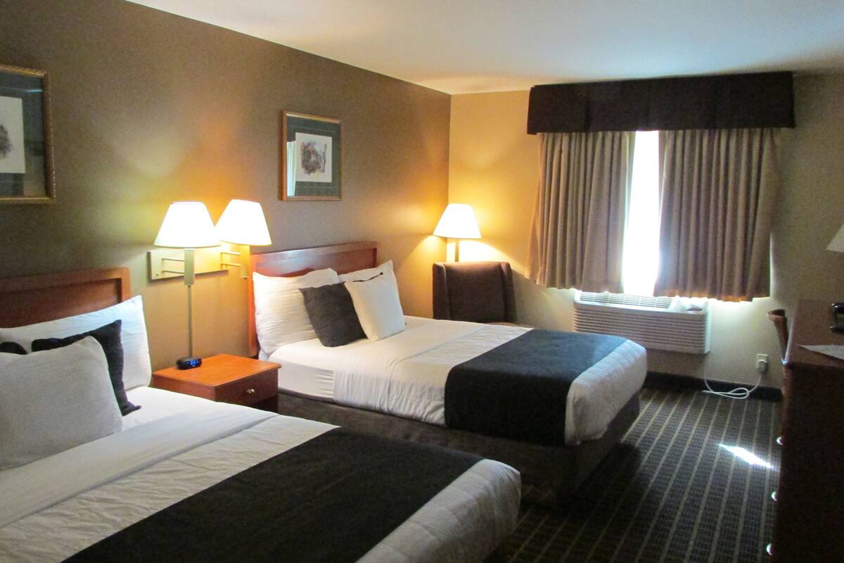 A hotel room with two beds, a nightstand, a lamp, a window with curtains, and a wall-mounted air conditioning unit.