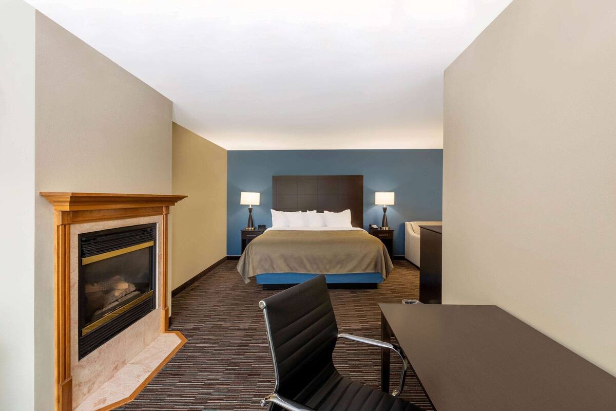 A hotel room with a bed, bedside lamps, a fireplace, a desk, and a chair.