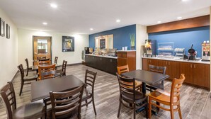 Comedor de hotel con mesas y sillas de madera, una encimera de cocina con una máquina de café y un bote de basura etiquetado como 'TRASH'.