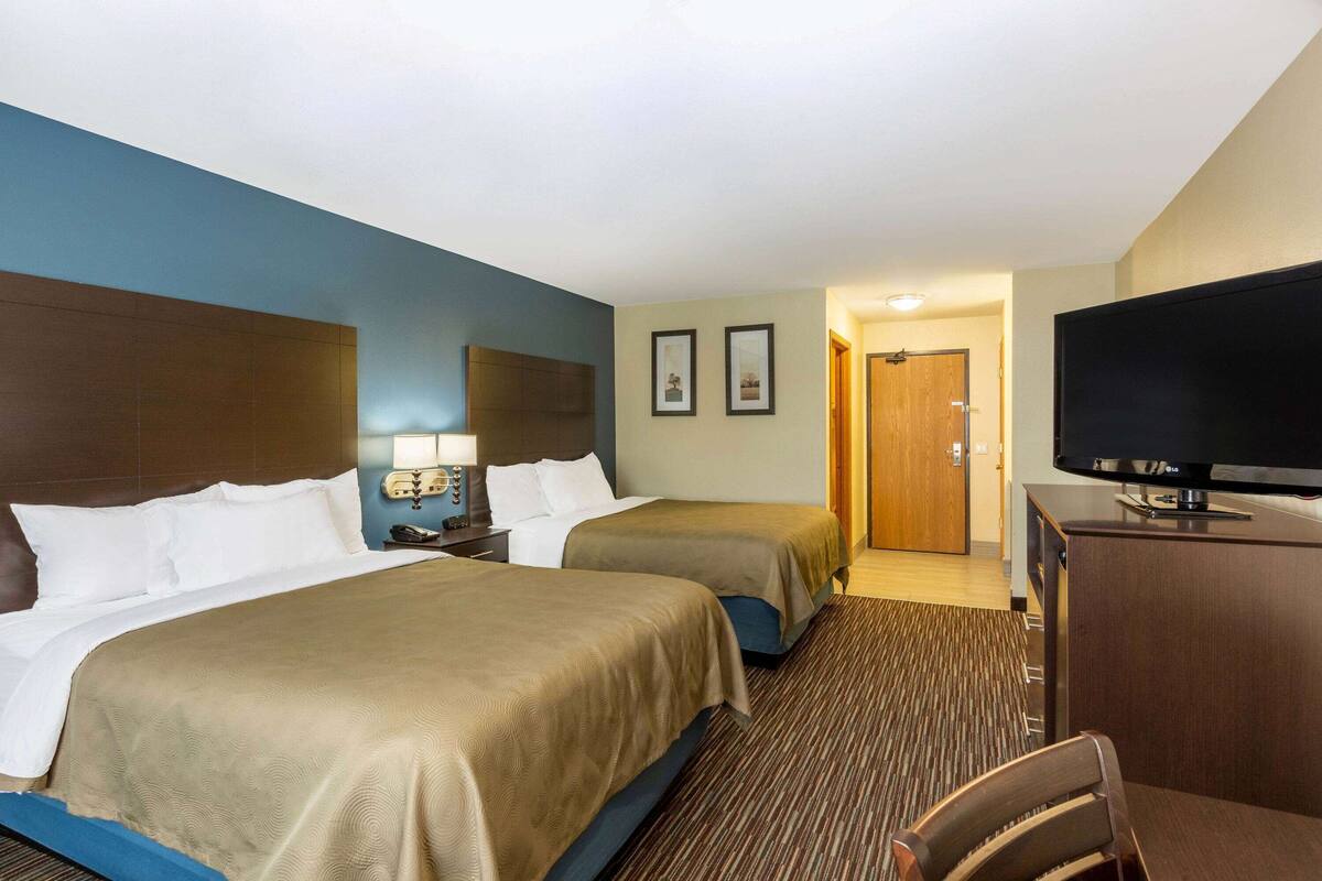 A hotel room with two beds, a desk with a flat-screen TV, a chair, a nightstand with a lamp, and a door leading to another room.