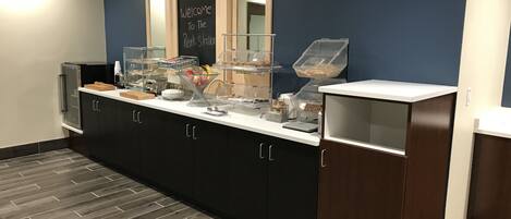 A breakfast bar with a chalkboard sign, a coffee machine, and various food containers.