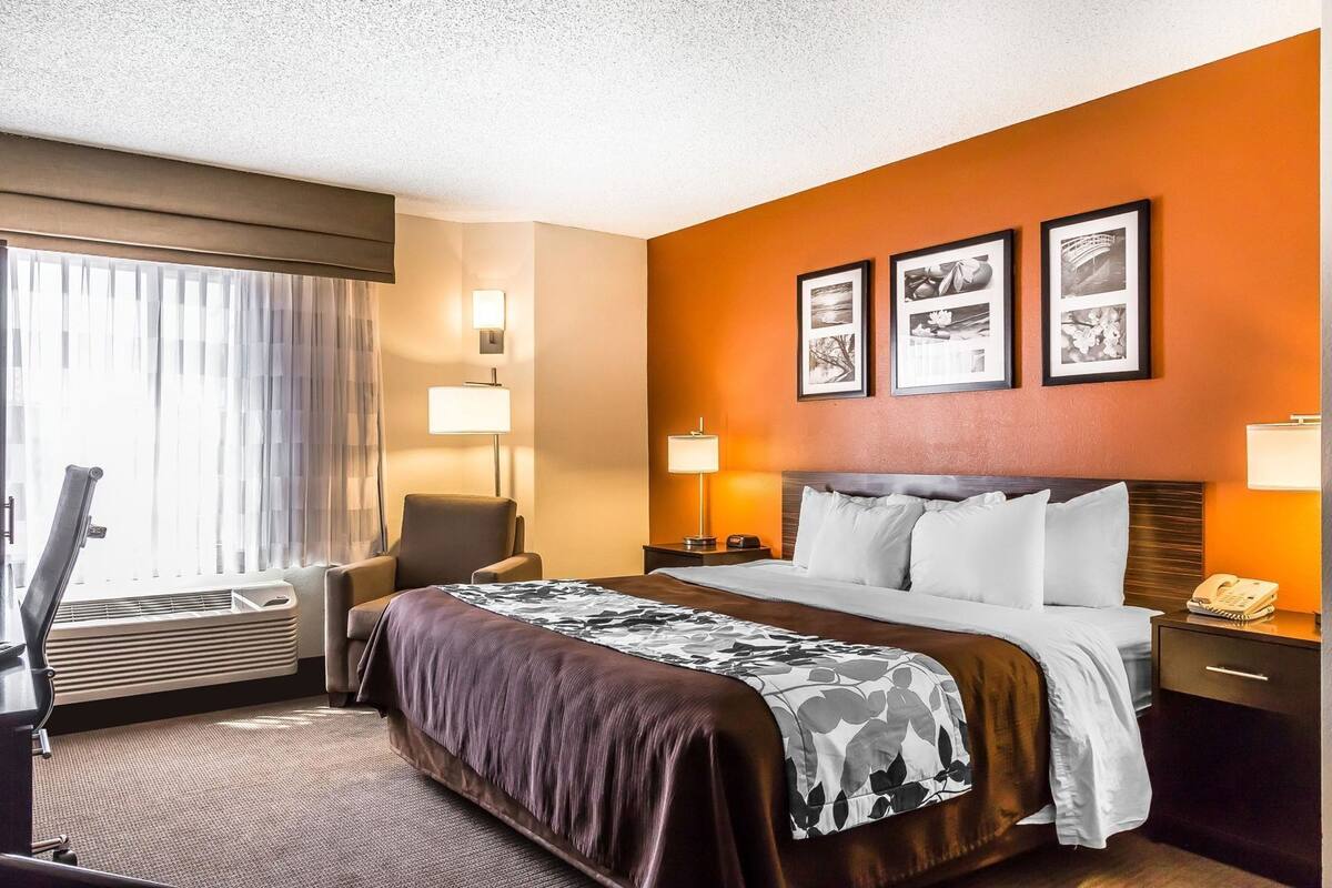 A hotel room with a bed, a chair, a desk, a lamp, a phone, and a window with curtains.