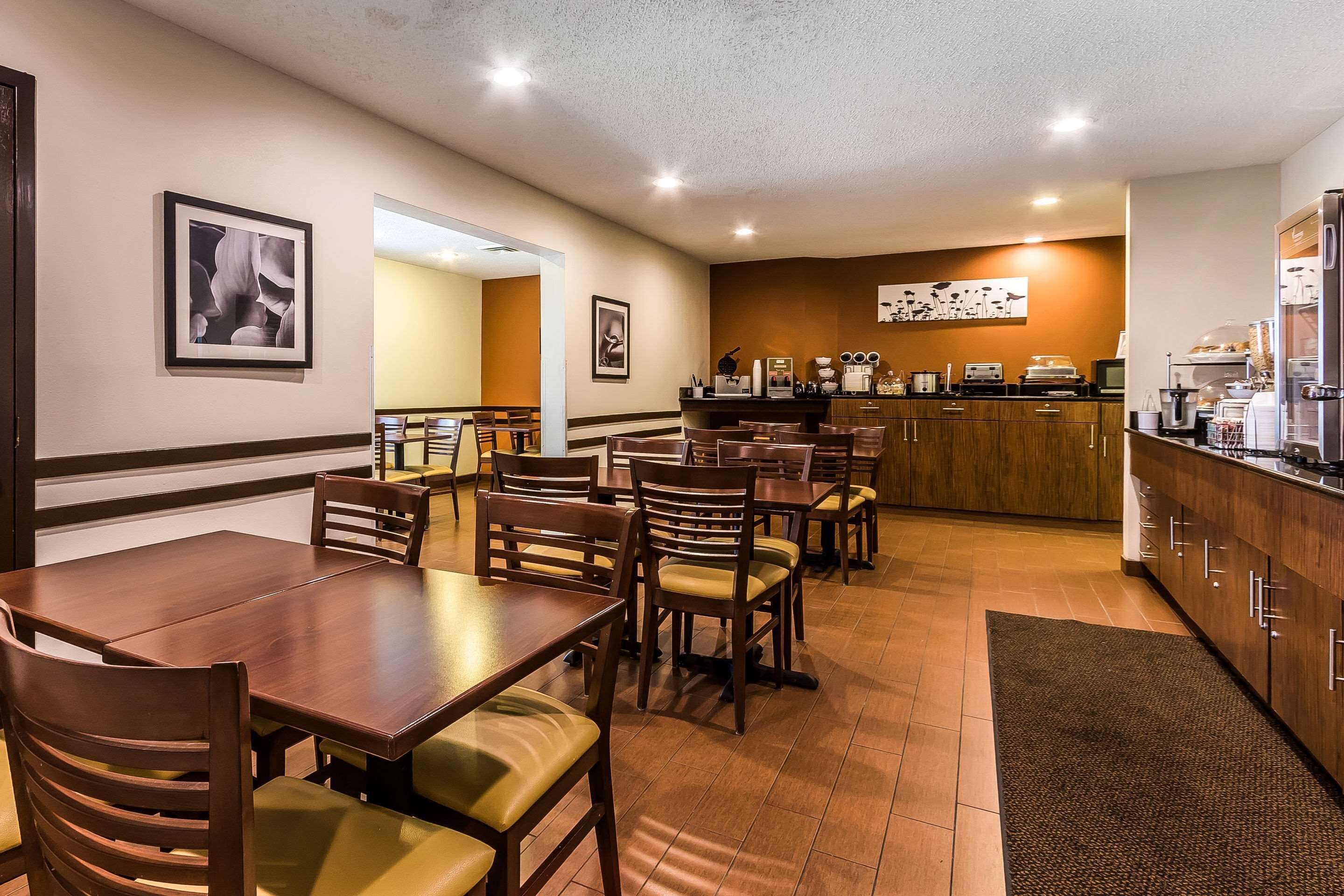 A hotel dining area with multiple wooden tables and chairs, a kitchen counter with appliances, and framed artwork on the walls.
