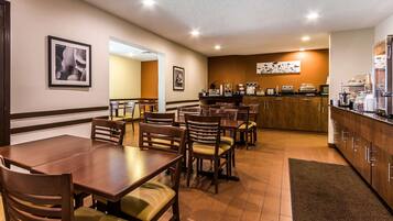 A hotel dining area with multiple wooden tables and chairs, a kitchen counter with appliances, and framed artwork on the walls.
