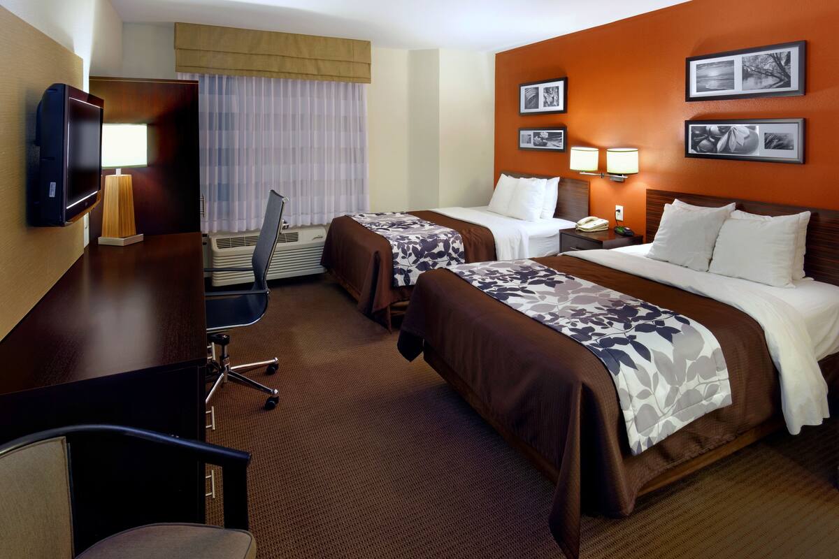 A hotel room with two beds, a desk with a chair, a TV, and a window with curtains.