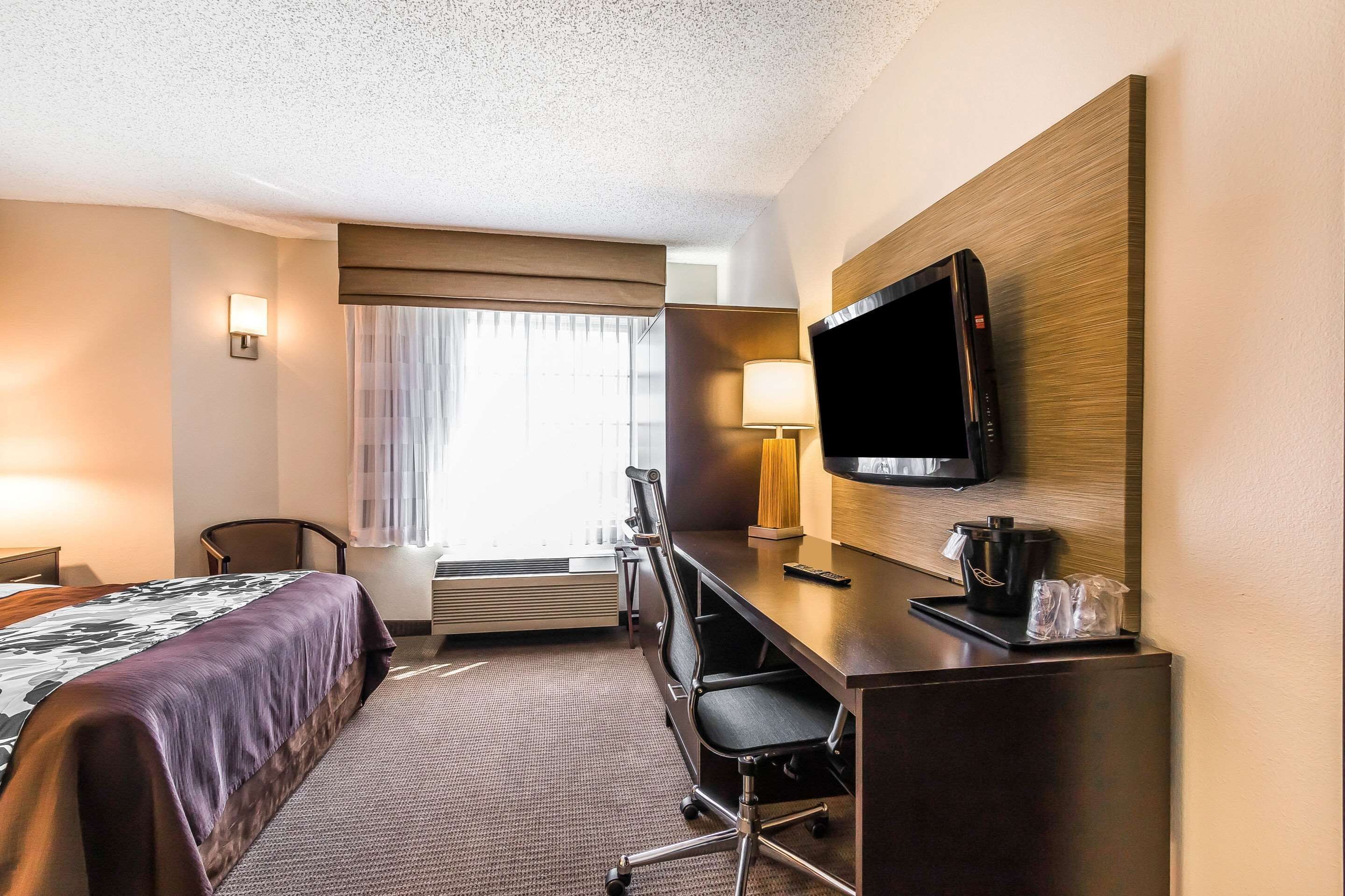 A hotel room with a bed, desk, chair, TV, lamp, and window with curtains.