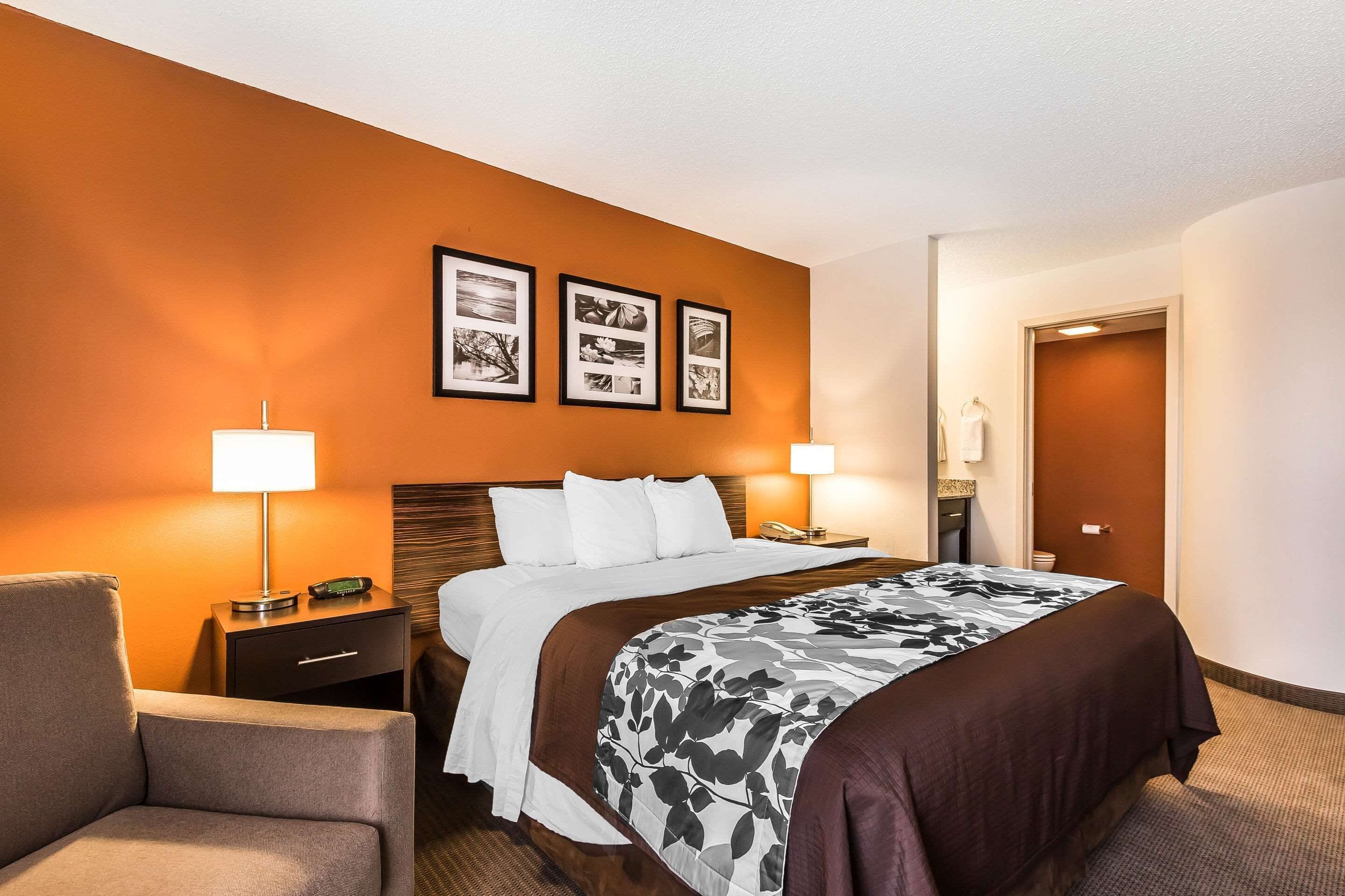 Quarto de hotel com uma cama grande, duas mesas de cabeceira com luminárias, uma cadeira e uma porta.