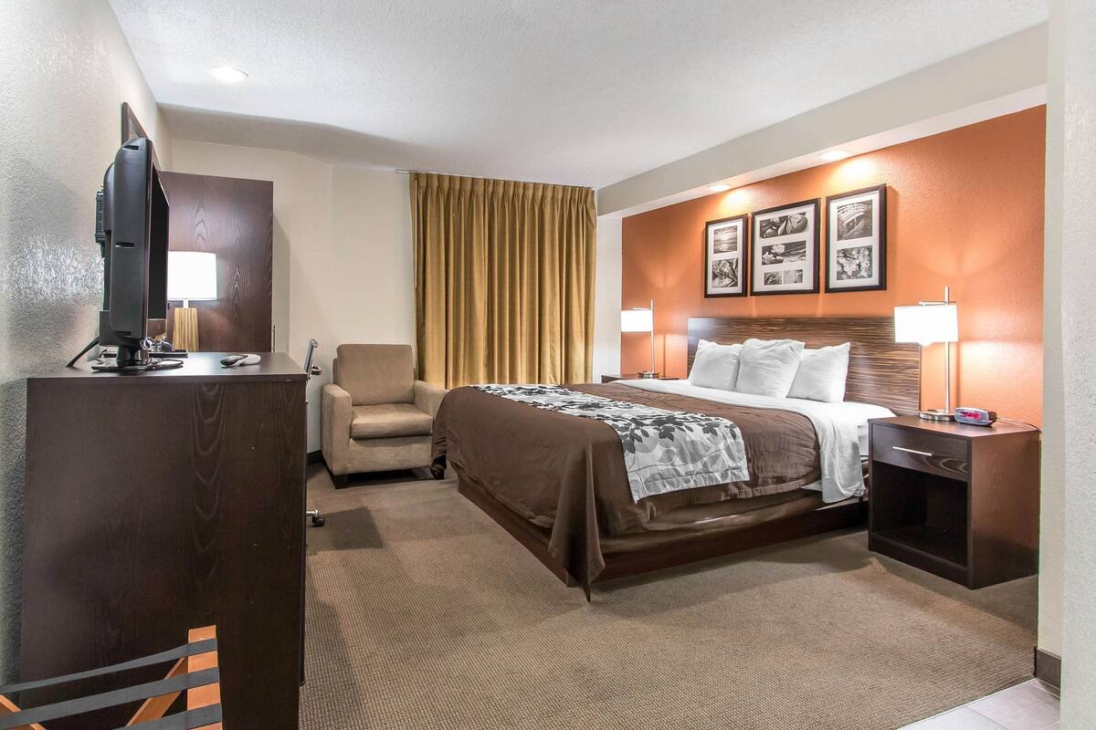 A hotel room with a bed, a desk with a computer, a chair, and a nightstand with a lamp.