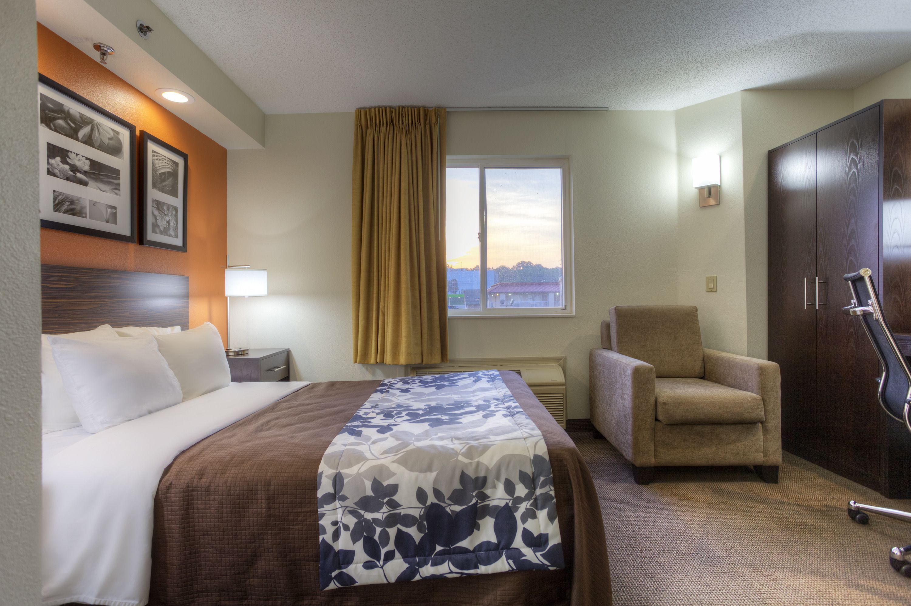 A hotel room with a bed, a chair, a desk, a lamp, and a window with a view.