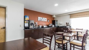 Uma área de refeições com mesas e cadeiras de madeira, cozinha com geladeira, cafeteira e bebedouro.