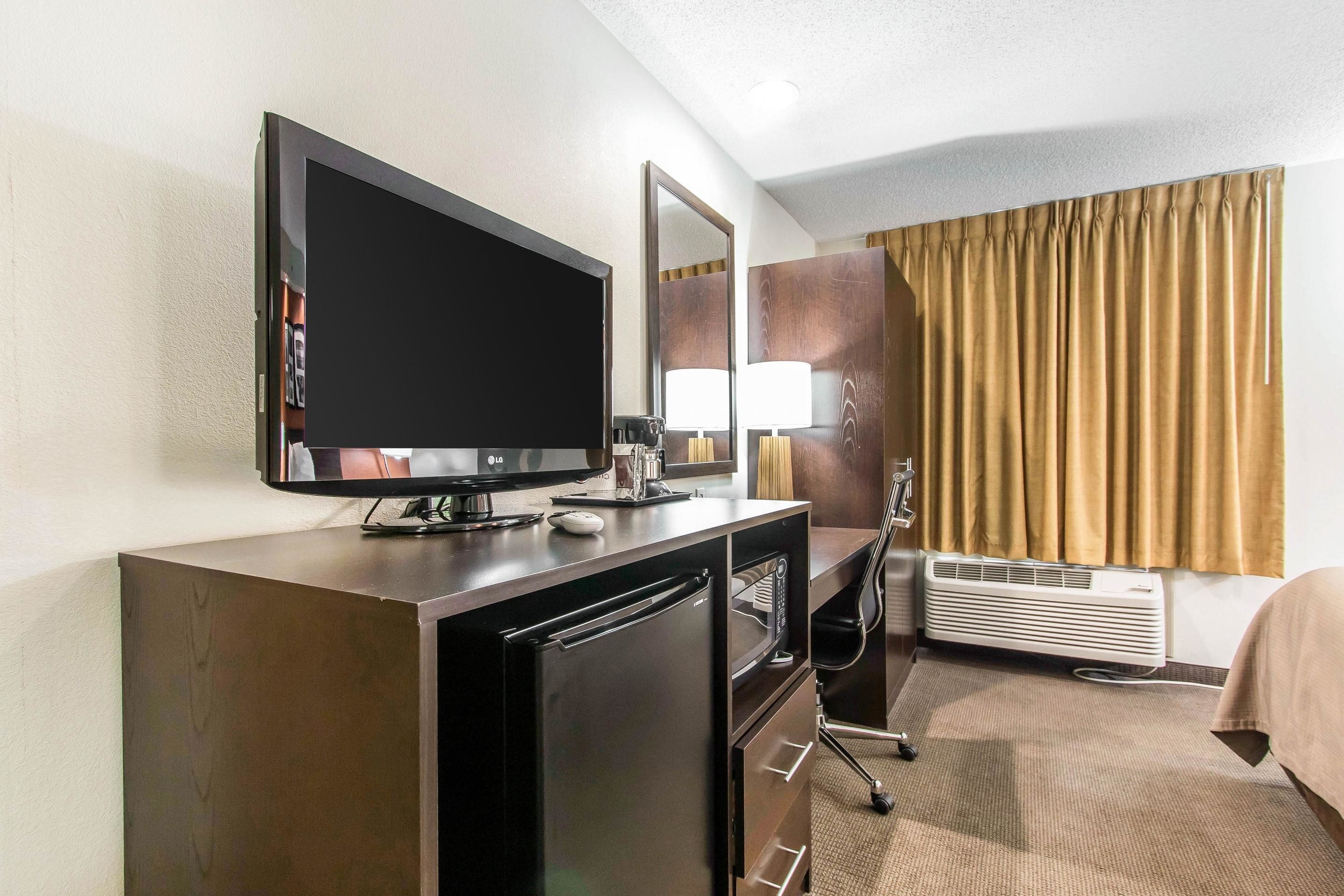 A hotel room with a flat-screen TV, a mirror, a desk with a lamp, a chair, an air conditioning unit, and a bed with a beige cover.