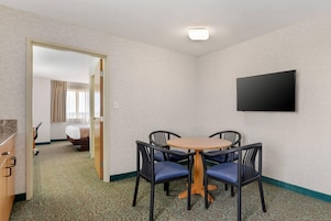 Habitación de hotel con cama, televisión montada en la pared, mesa de comedor con sillas y piso alfombrado.