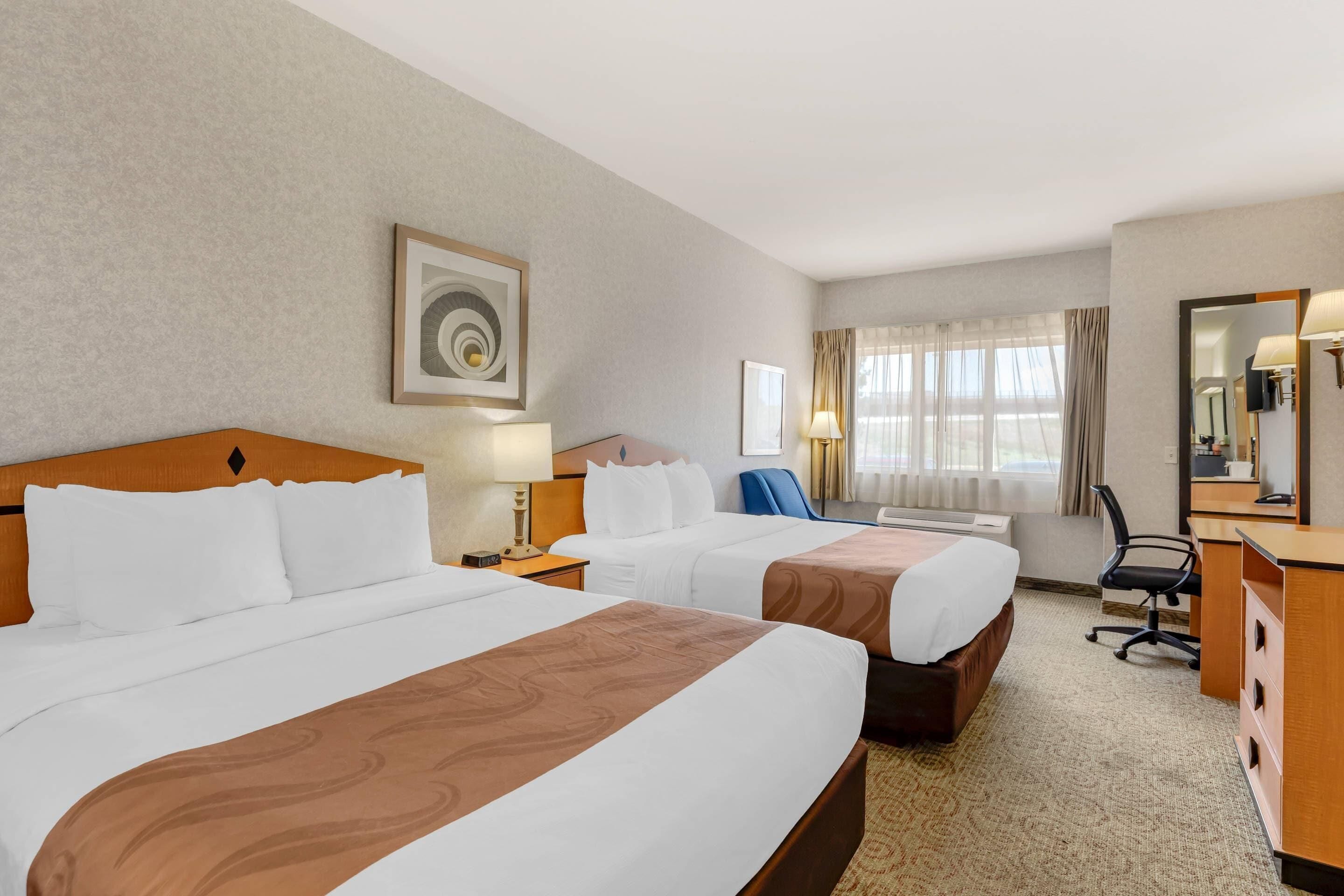 Habitación de hotel con dos camas, un escritorio, una silla y un ventanal con cortinas.