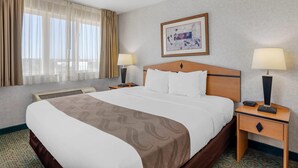 Una habitación de hotel con una cama grande, dos mesitas de noche con lámparas, una ventana con cortinas, un cuadro en la pared y suelo alfombrado.