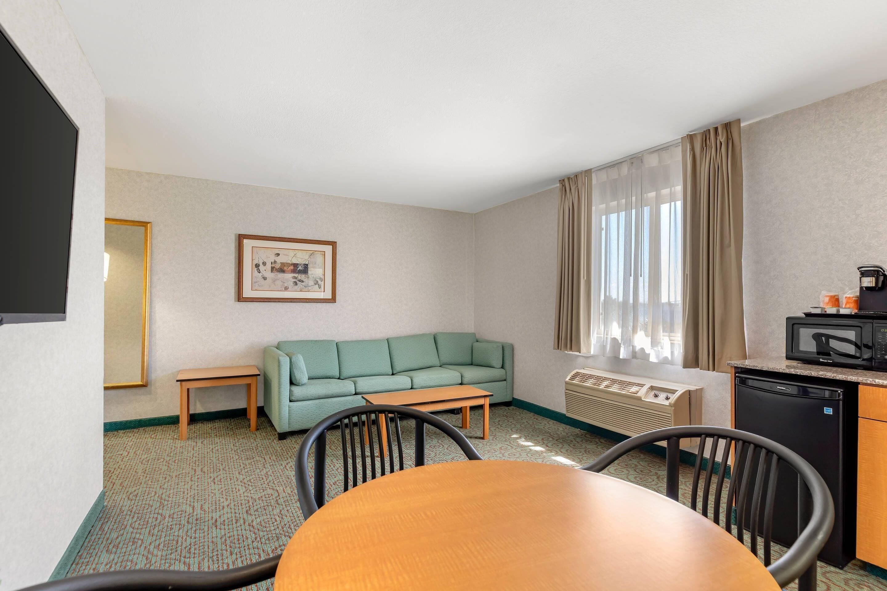 A hotel room with a green sofa, a wooden table, a dining area, a kitchenette, and a window with curtains.
