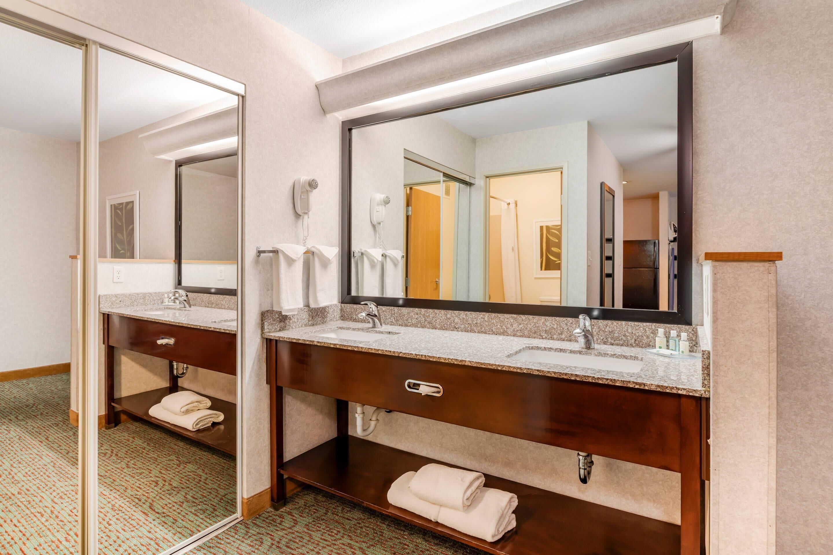 A hotel bathroom with two sinks, a mirror, a hairdryer, and a towel rack.