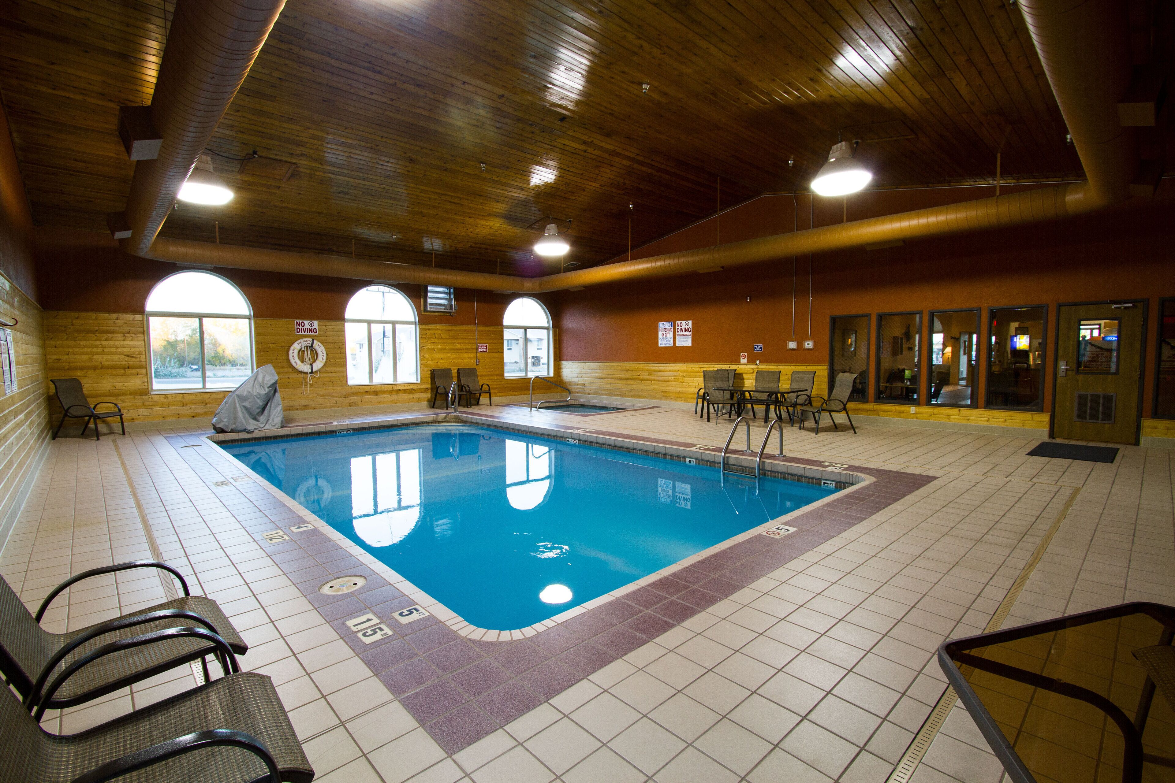 An indoor swimming pool with clear lane markings, surrounded by lounge chairs and tables, and large windows providing natural light.
