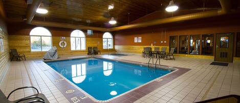 An indoor swimming pool with clear lane markings, surrounded by lounge chairs and tables, and large windows providing natural light.