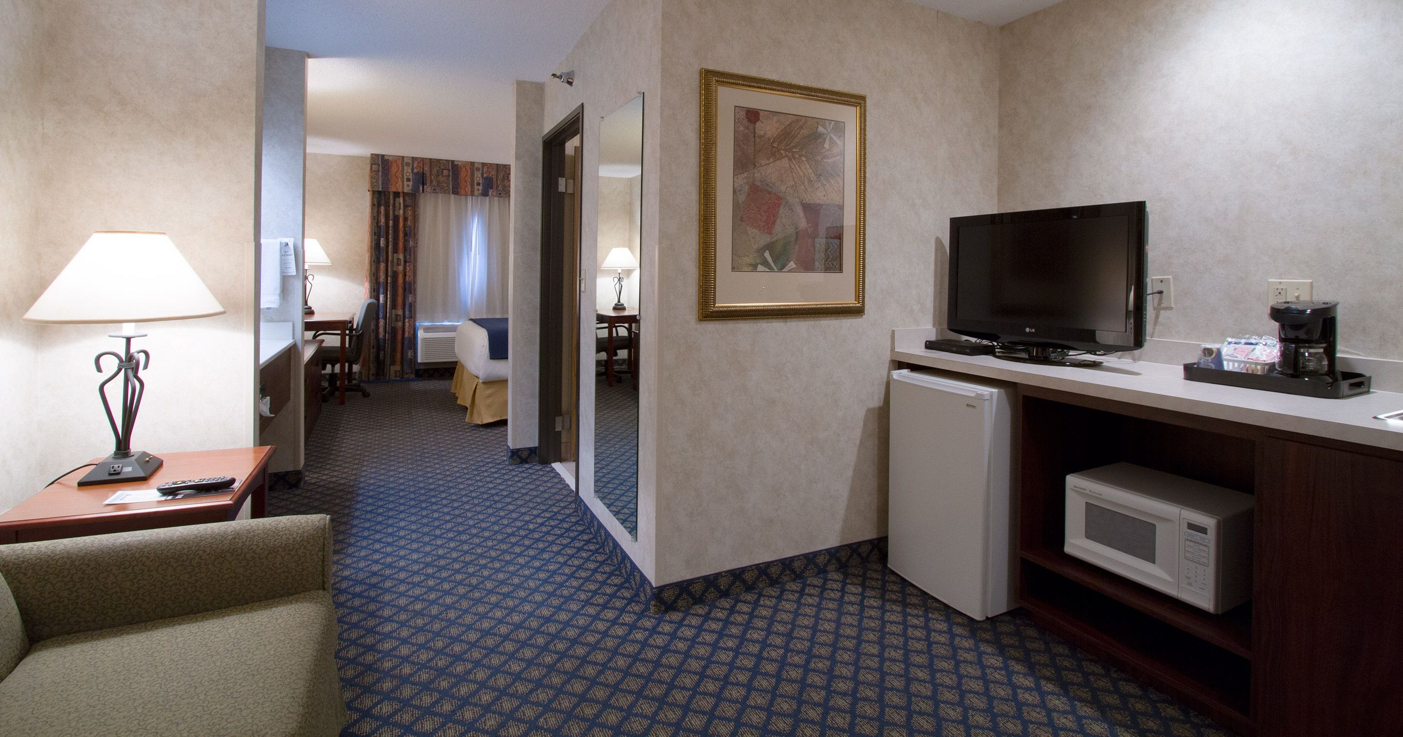 A hotel room with a bed, a desk with a lamp, a television, a microwave, and a refrigerator.