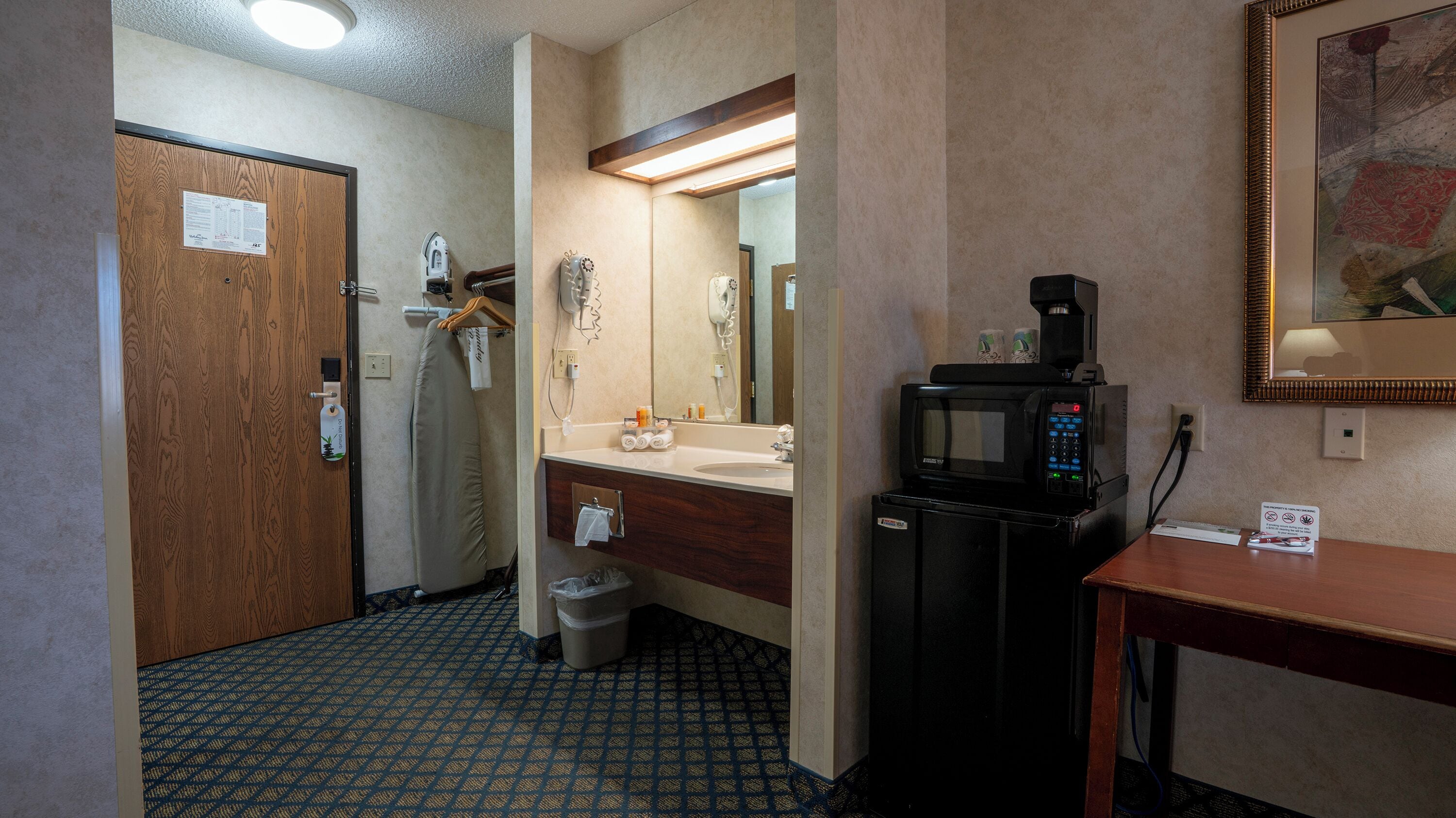Une salle de bain d’hôtel avec une porte en bois, un lavabo, un miroir, un sèche-cheveux, une poubelle et une machine à café.