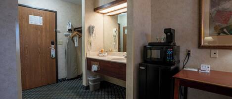 Une salle de bain d’hôtel avec une porte en bois, un lavabo, un miroir, un sèche-cheveux, une poubelle et une machine à café.