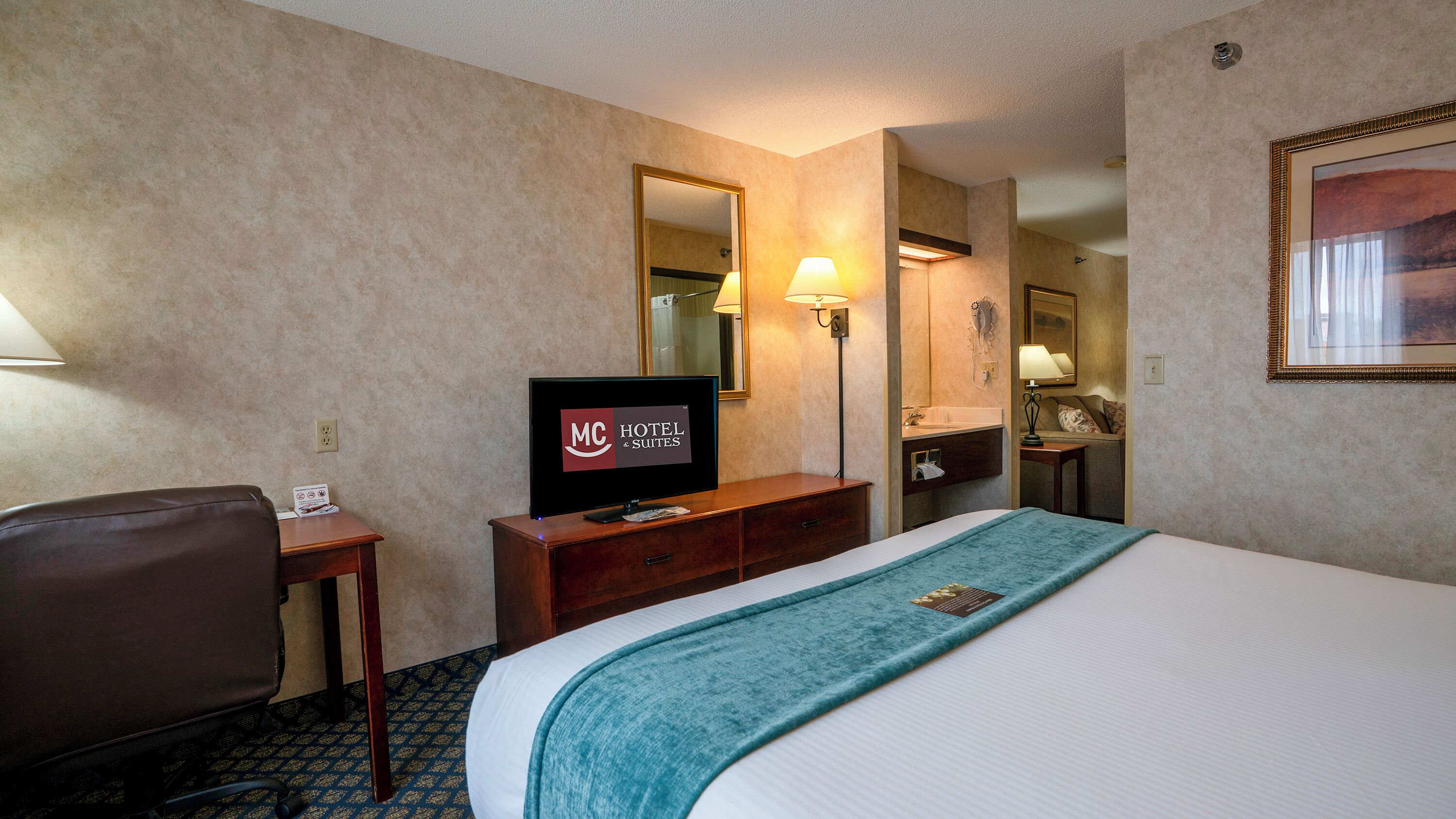 A hotel room with a bed, a desk with a television displaying the hotel name, a chair, a nightstand, a mirror, and a bathroom visible through an open door.
