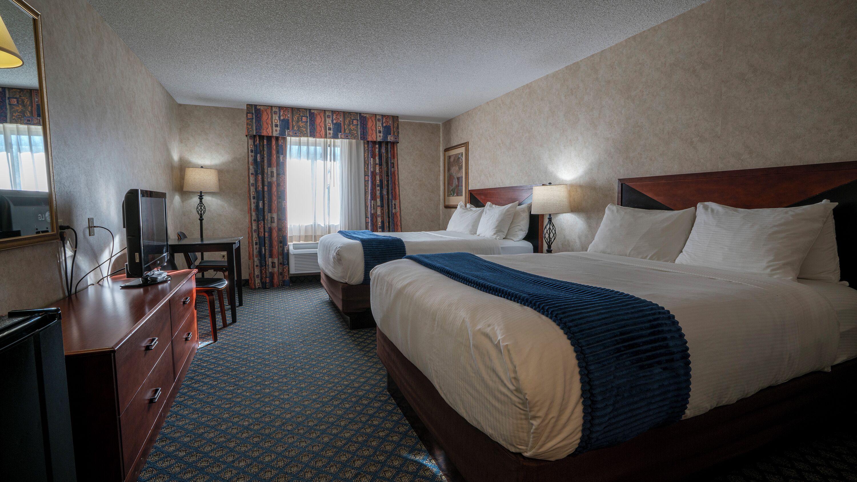 A hotel room with two beds, a desk with a computer, a TV, and a window with curtains.