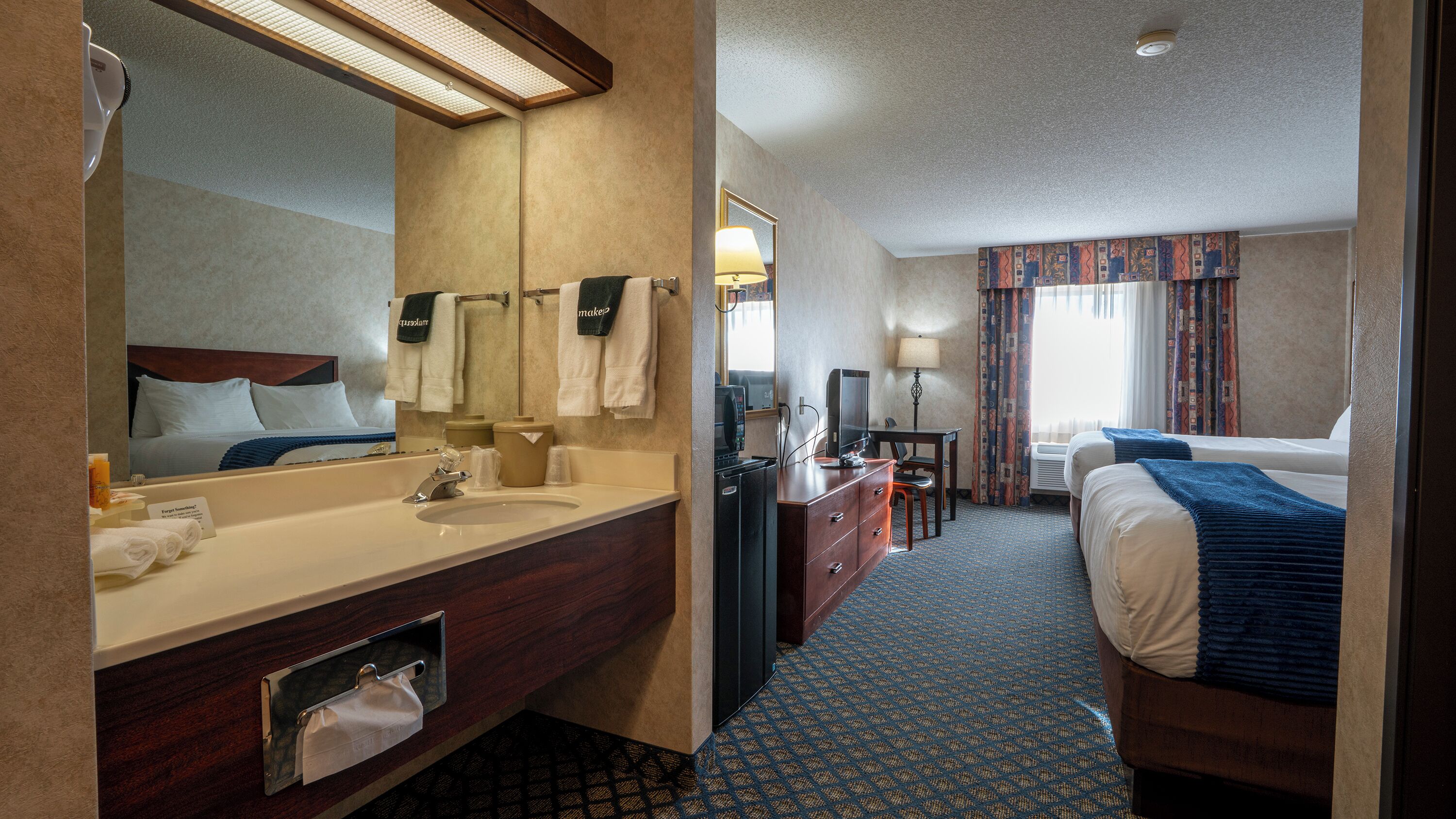 Habitación de hotel con un espejo grande, un lavamanos, un escritorio con computadora, una cama y una ventana con cortinas.