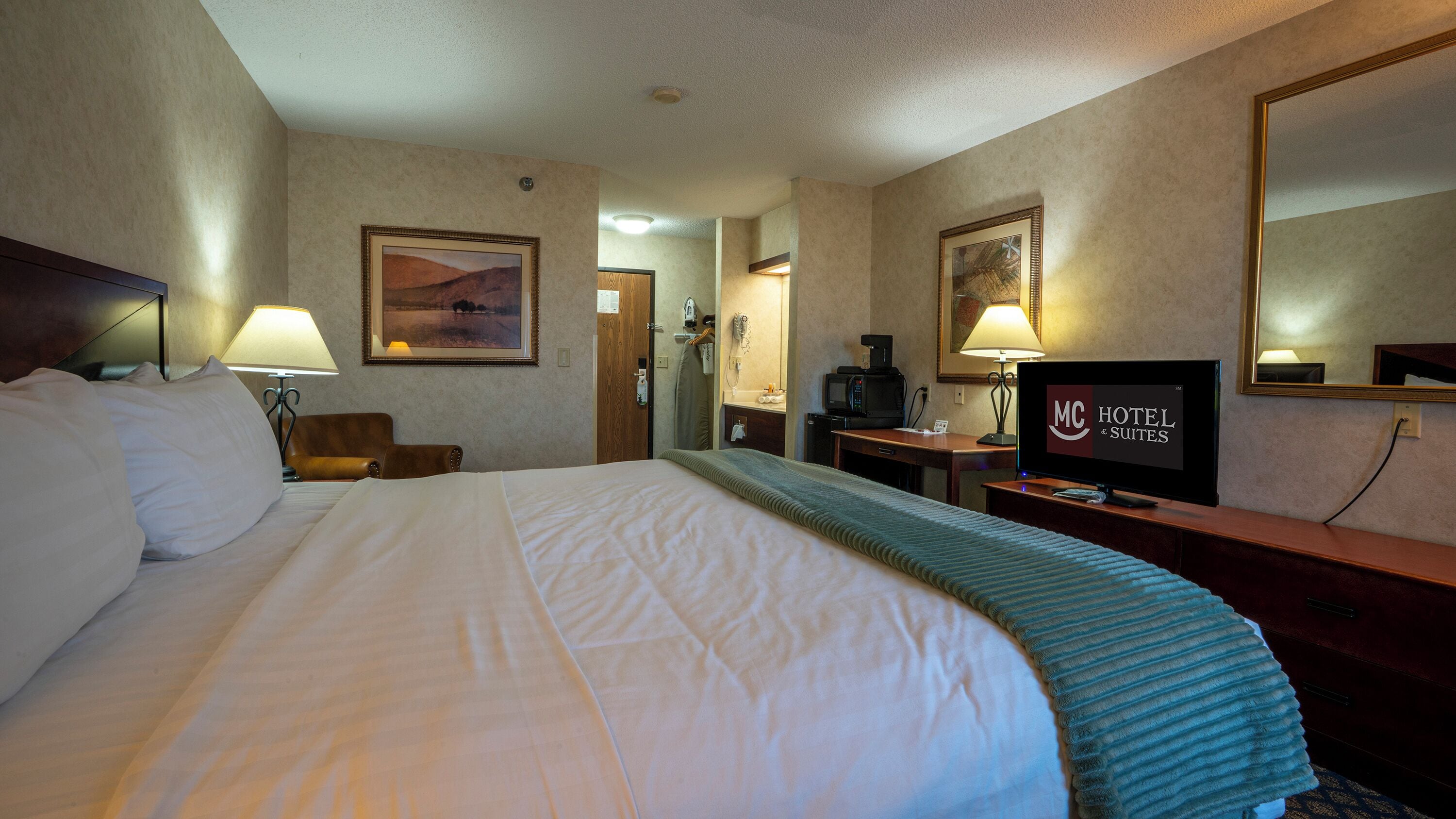 Habitación de hotel con una cama grande, un escritorio con televisor, un espejo y un cuadro en la pared.