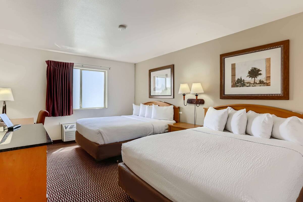 A hotel room with two beds, a desk, a chair, a window with curtains, a wall-mounted mirror, and a framed picture.