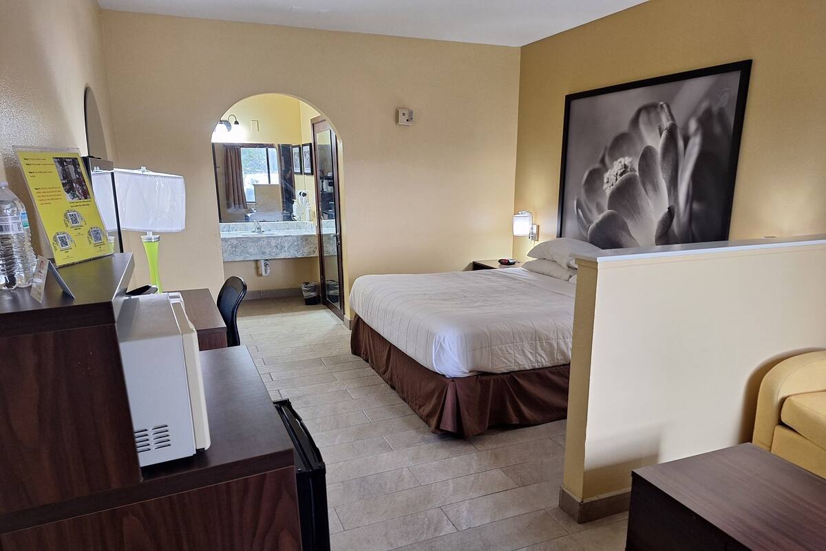 A hotel room with a bed, a desk with a computer, a chair, a mirror, and a framed picture on the wall.