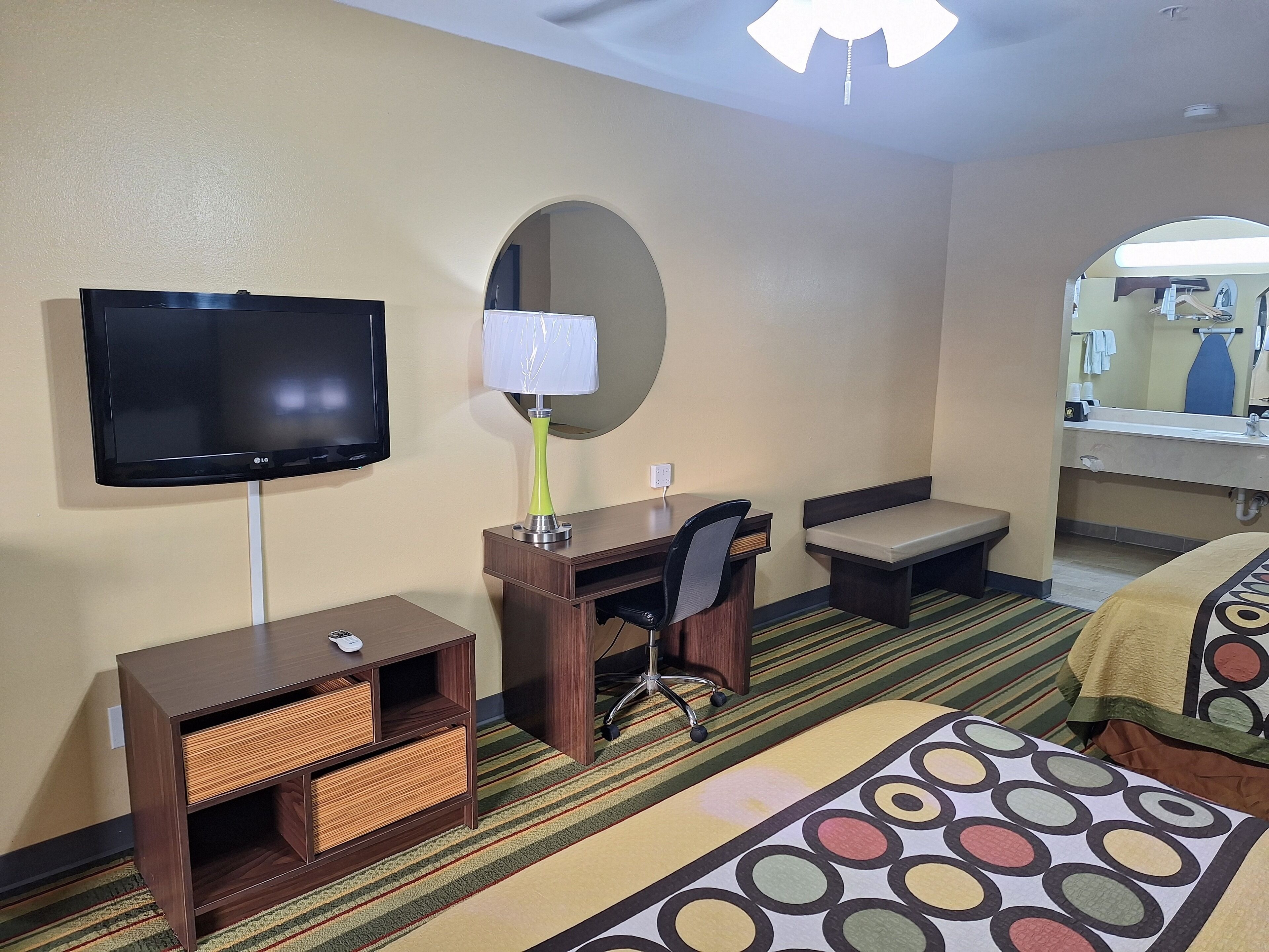 A hotel room with a television, desk, chair, bench, and a patterned rug.