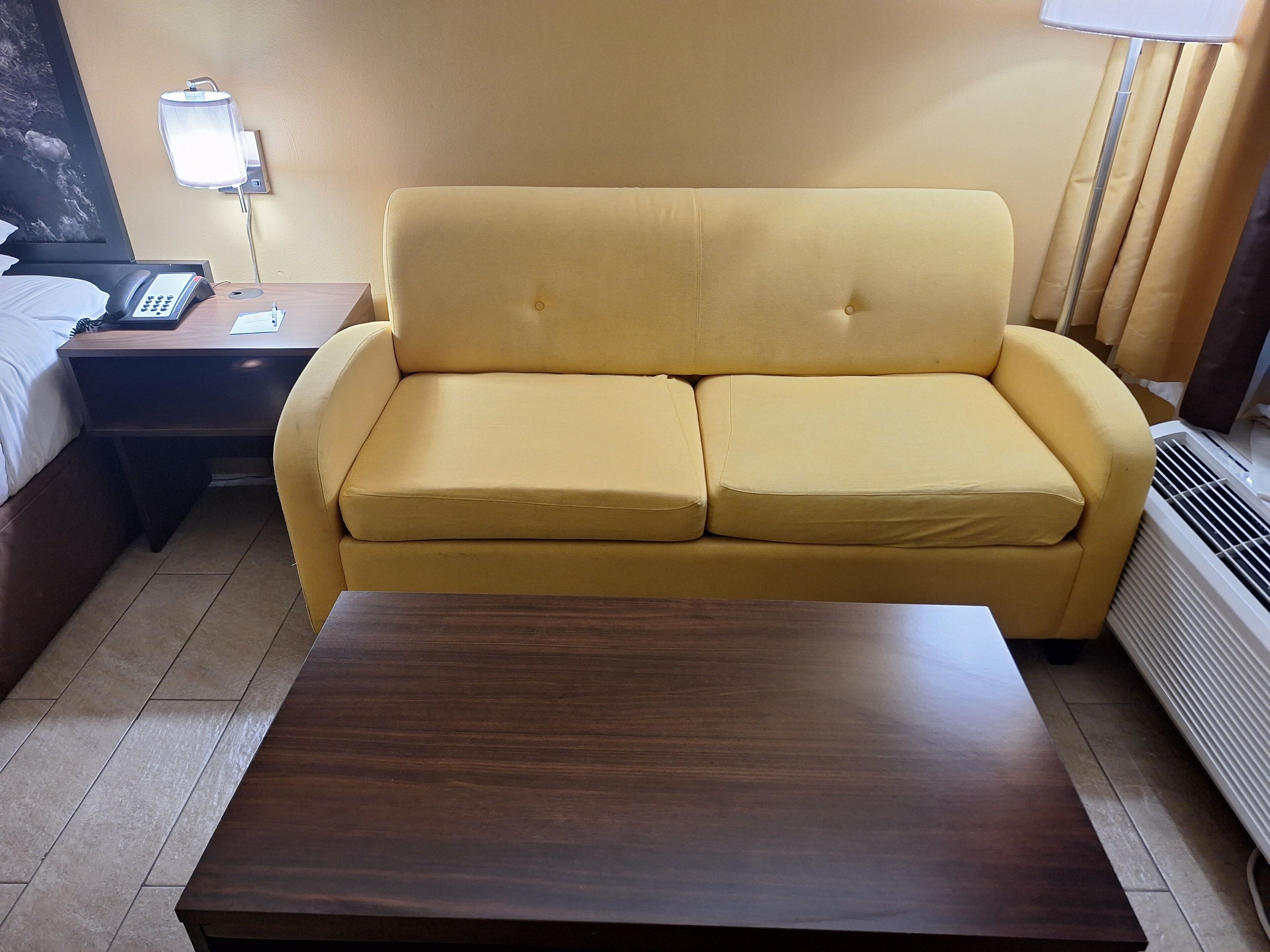 A yellow sofa in a hotel room with a wooden side table, a bedside phone, and a wall-mounted lamp.
