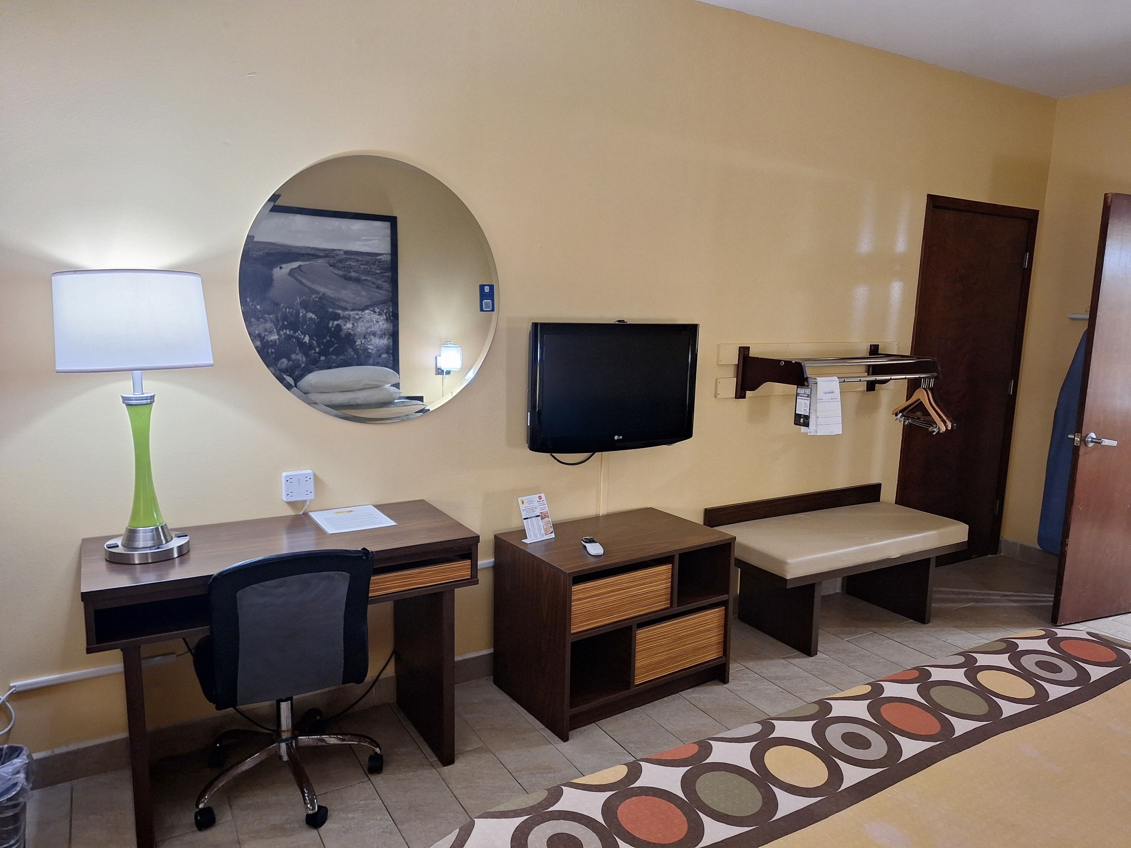 A hotel room with a desk, chair, TV, bedside table, and a bench.