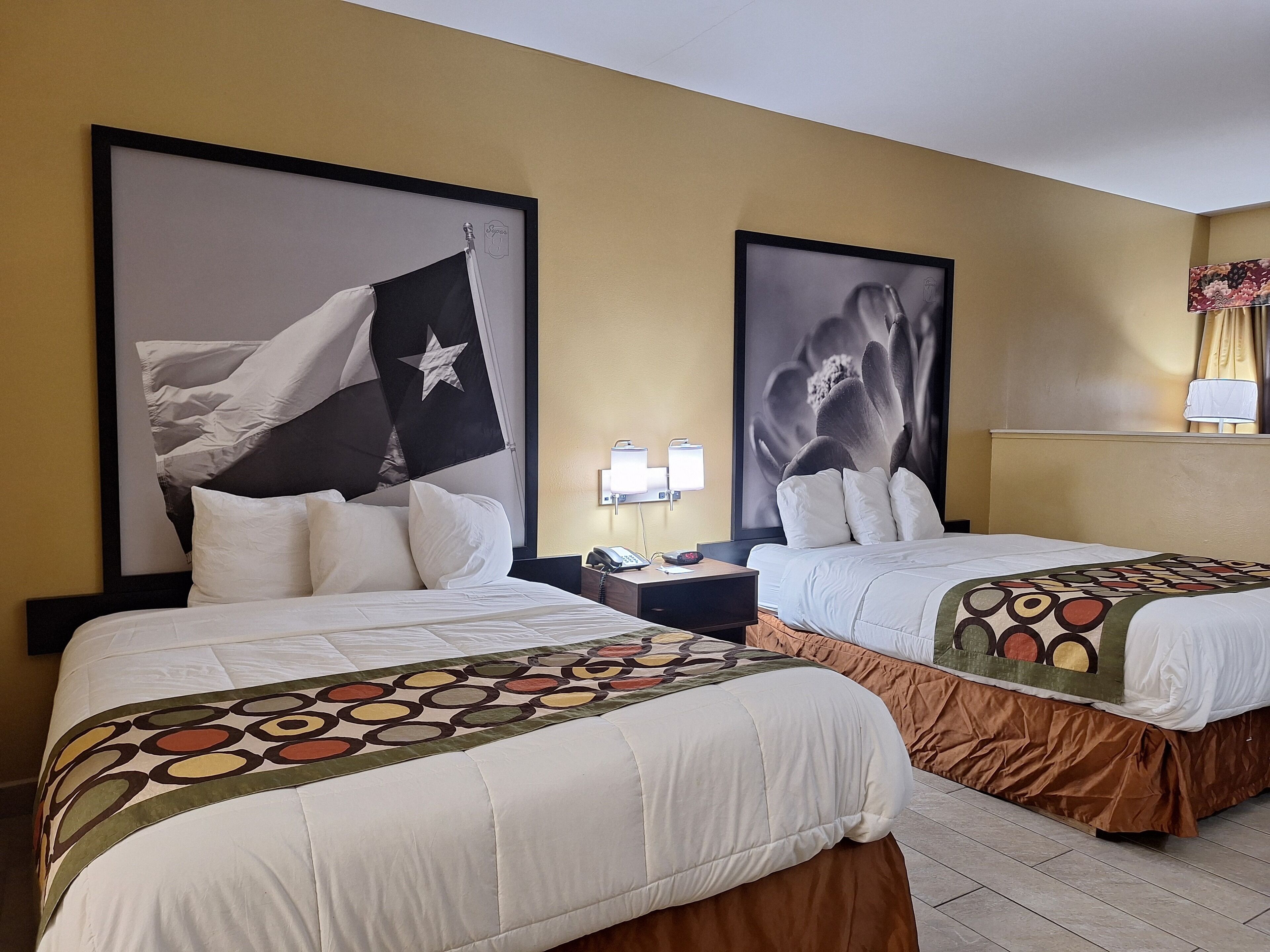 A hotel room with two beds, each with white bedding and a patterned throw. There are two nightstands with lamps and a phone. The walls are painted yellow, and there are two large framed pictures above the beds.