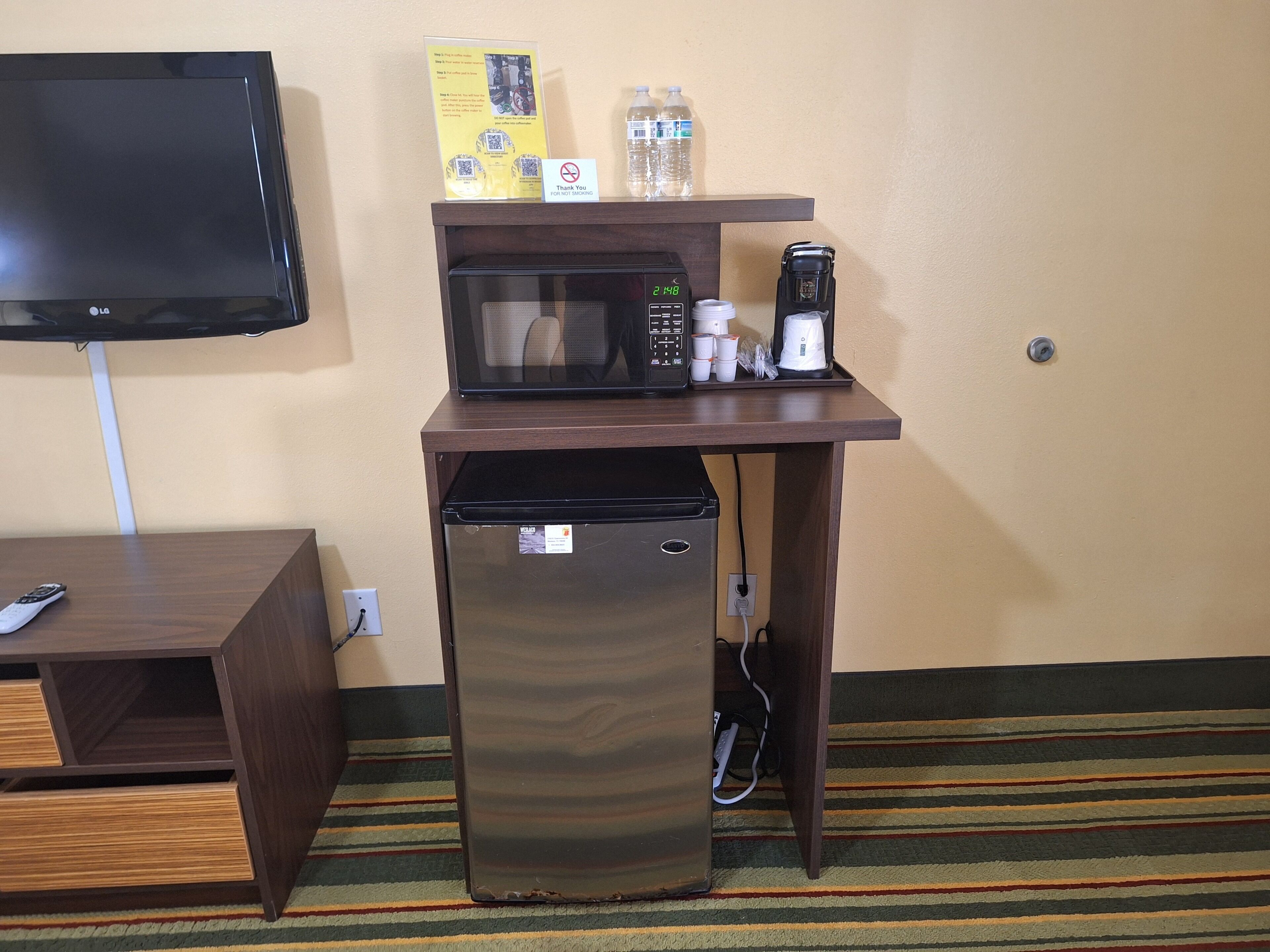 A compact kitchenette with a microwave, coffee maker, and a refrigerator. A TV is mounted on the wall above the kitchenette.