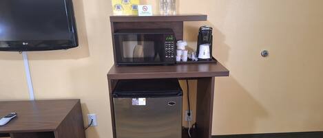 Una kitchenette compacta con microondas, cafetera y refrigerador. Un televisor está montado en la pared sobre la kitchenette.