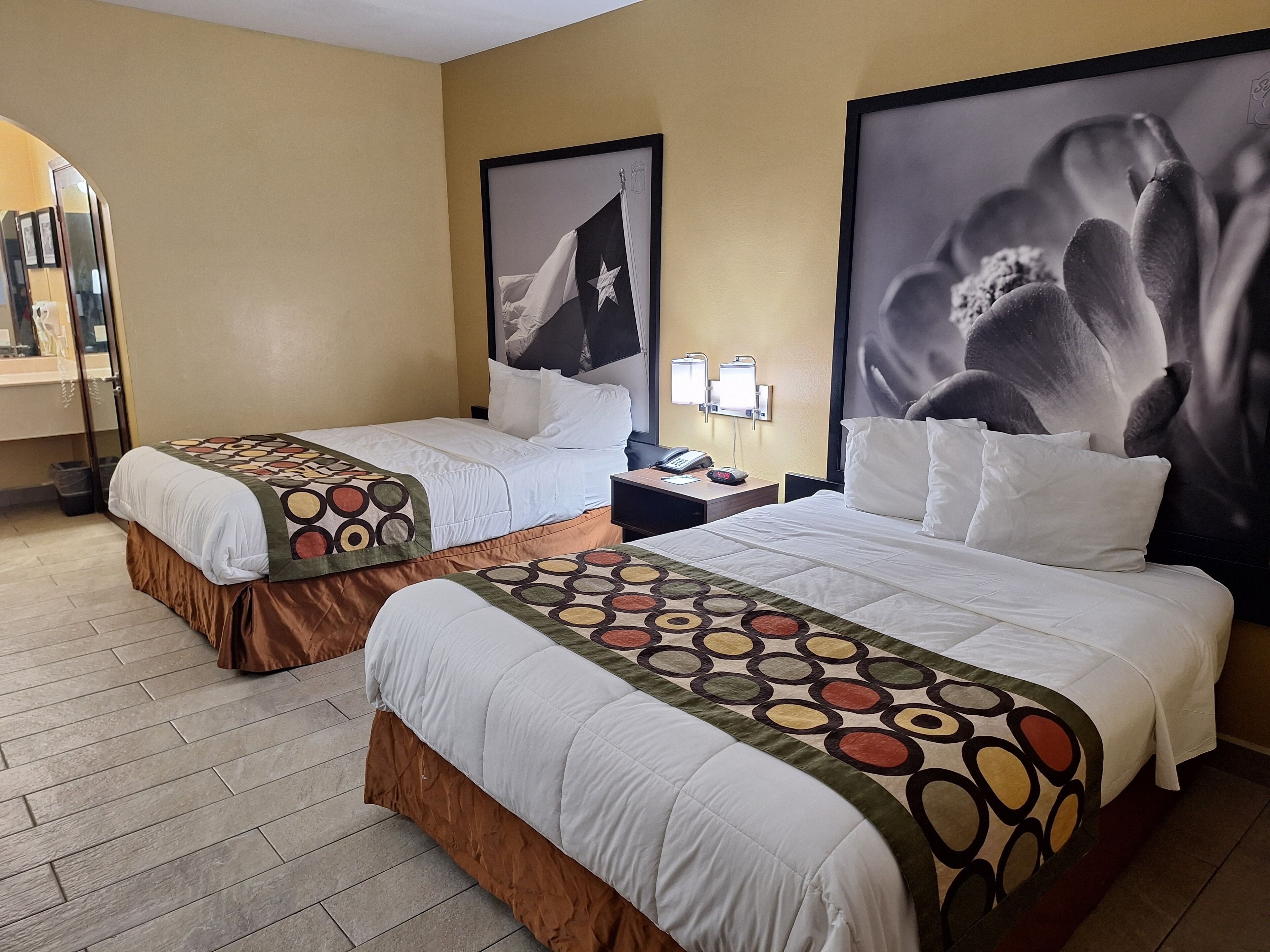 A hotel room with two beds, each with a patterned bedspread and a brown skirted bed base. There is a nightstand with a phone and a lamp between the beds. The walls are beige, and there are two large framed pictures above the beds.