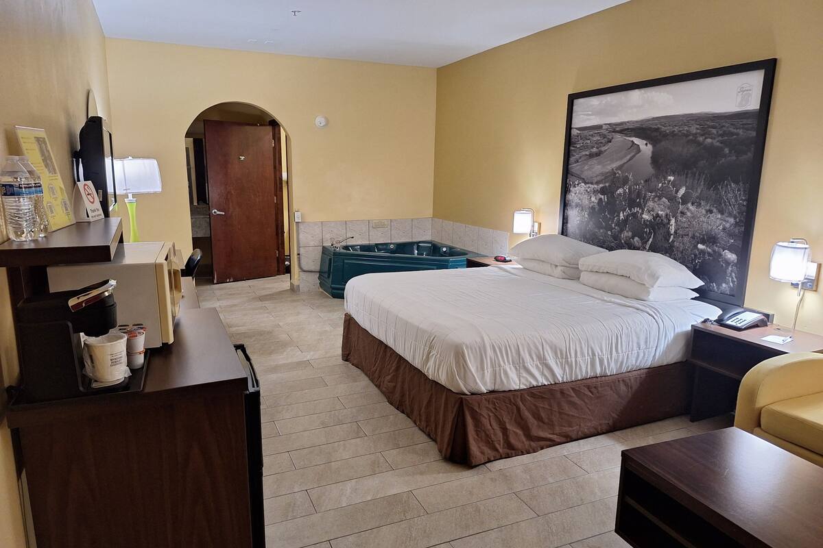 A hotel room with a large bed, a desk with a computer, a desk lamp, a telephone, a large framed picture on the wall, and a bathtub.